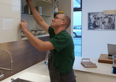 Ein Mann in grünem T-Shirt, welcher an einem Küchenschrank werkelt und dabei lächelt.