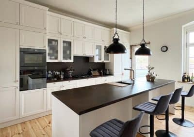 Eine weiße Küchenzeile mit schwarzer Oberfläche, an welcher Barhocker stehen. Im Hintergrund ein großer, weißer Küchenschrank mit einer Vielzahl an Schubladen und Türen.