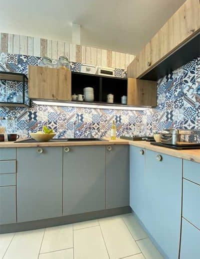 Eine Küche mit olivgrüner Verkleidung und hellen Holzplatten als Oberfläche. An der Wand verschieden gemusterte, hauptsächlich blaue Fliesen.