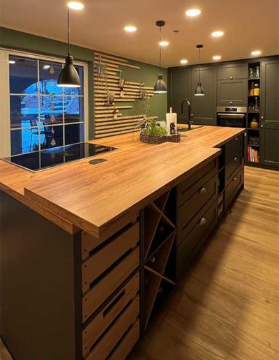 Eine schwarze Küchenzeile mit heller Holzplatte als Oberfläche, auf welcher eine Herdplatte aufliegt. Im Hintergrund schwarze Küchenschränke und links eine olivgrüne Wand mit Holzbrettern.