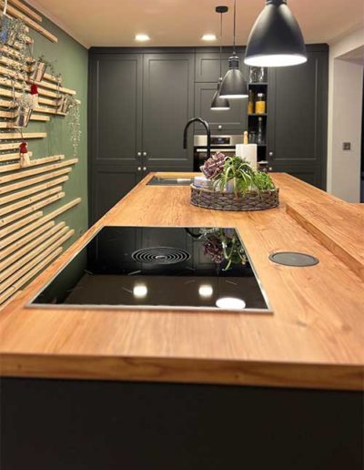 Eine schwarze Küchenzeile mit heller Holzplatte als Oberfläche, auf welcher eine Herdplatte aufliegt. Im Hintergrund schwarze Küchenschränke und links eine olivgrüne Wand mit Holzbrettern.