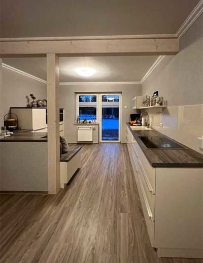 Blick auf eine Küche mit einer langgezogenen Küchenzeile rechts und einigen Küchenschränken links, welche von einem Holzbalken mittig abgetrennt werden.