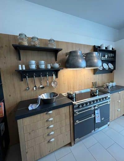 Eine Küche mit teils hölzernen Küchenschränken und einem schwarzem Herd. Darüber zwei prägnante Stahllampen.
