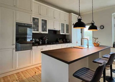 Eine weiße Küchenzeile mit schwarzer Oberfläche auf Holzboden, an welche Barhocker gestellt sind. Im Hintergrund eine große weiße Holzküche.