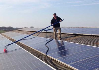 Ein Mann auf einem mit Photovoltaikplatten bedecktem Dach, welcher die Platten mit einer langen elektrischen Reinigungsbürste reinigt.