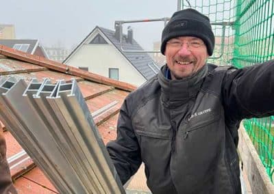 Ein Mann in Arbeitskleidung auf einem Gerüst an einem Dach, welcher in die Kamera lächelt.