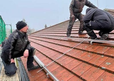 Drei Männer in Arbeitskleidung, welche Stahlträger für Photovoltaikplatten auf einem Dach anbringen.
