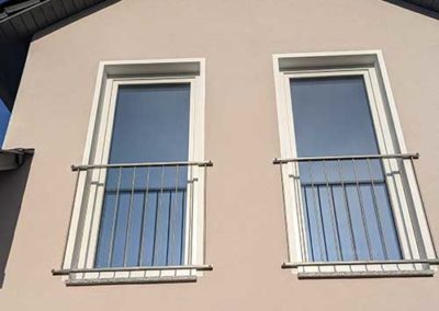 Die Fassade eines Hauses mit zwei Fenstern, an welchen stählerne Geländer angebracht sind.