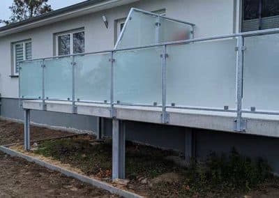 Stahlgeländer mit Glasplatten als Geländer eines Balkons eines Wohnhauses. Der Balkon ist über Stahlgerüste mit dem Boden verbunden.