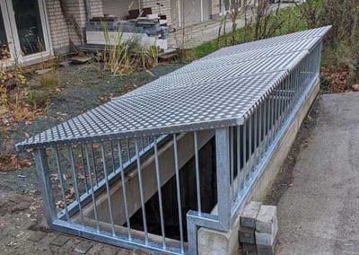 Ein stählernes Gitter mit Stahldach, welches eine Öffnung im Boden vor einem geklinkerten Wohnhaus abdeckt.